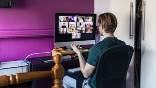 [Translate to English:] Blick über die Schulter: Ein Studierender sitzt zu Hause vor dem PC und blickt auf eine Videokonferenz auf dem Bildschirm, der vor einer lila Wand steht. Foto: © SolStock/ E+/ Getty Images