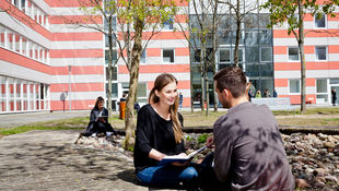 Zwei Studierende sitzen am Campus Lichtenberg im Hof unter einem Baum und unterhalten sich.