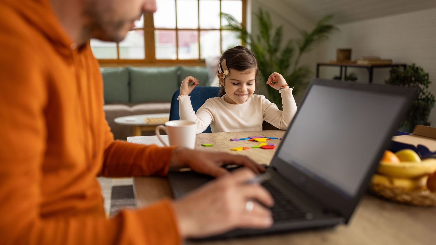 Mann arbeitet am Laptop, im Hintergrund sitzt ein Kind und malt