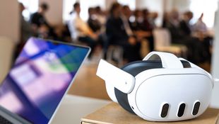 Laptop und VR Brille liegen auf einem Tisch, Menschen im Hintergrund
