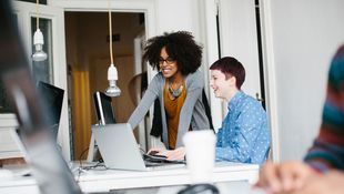 Zwei Frauen arbeiten vor einem Schreibtisch mit Computer und Laptop.