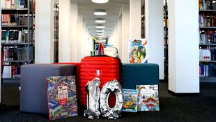 Roter Koffer mit silbernen Luftballons in Form der Zahl 10 und mehreren Büchern vor Bücherregalen in einem hellen Raum