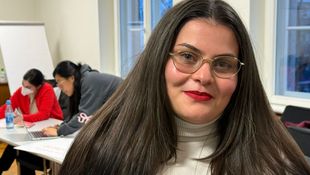 Junge Frau mit langem dunklem Haar und hellem Rollkragenpullover in einem Raum mit mehreren Personen und Flipchart im Hintergrund