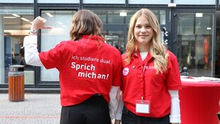 Zwei Frauen in roten Poloshirts stehen nebeneinander, eine zeigt mit dem Daumen auf den Rücken ihres Shirts mit der Aufschrift 'Ich studiere dual. Sprich mich an!'