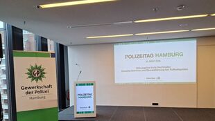 Konferenzraum mit Leinwand, auf der 'POLIZEITAG HAMBURG 28. MÄRZ 2023 Störungseffekte trotz Hochrisiko Gewaltprävention und Einsatzführung bei Fußballspielen' steht, daneben ein Banner und ein Pult mit dem Logo der Gewerkschaft der Polizei Hamburg