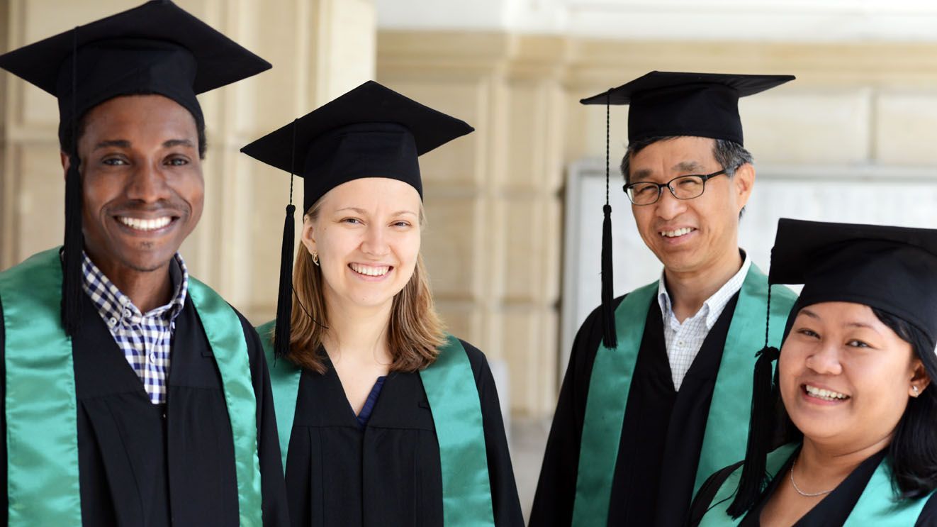 Vier Personen in schwarzen Talaren mit grünen Schärpen und schwarzen Doktorhüten stehen nebeneinander vor einer hellen Wand.