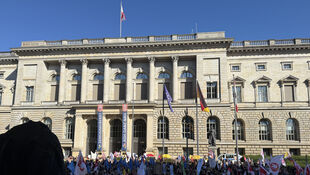 Menschenmenge mit Protestschildern vor einem historischen Gebäude mit Säulen und Flaggen