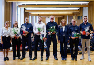 Sieben Personen stehen nebeneinander in einem Saal, einige tragen Uniformen, andere halten Blumen und Geschenke in den Händen.