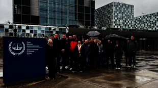 Gruppe von Menschen mit Regenschirmen vor dem Gebäude des Internationalen Strafgerichtshofs mit blauem Schild und Waage-Logo